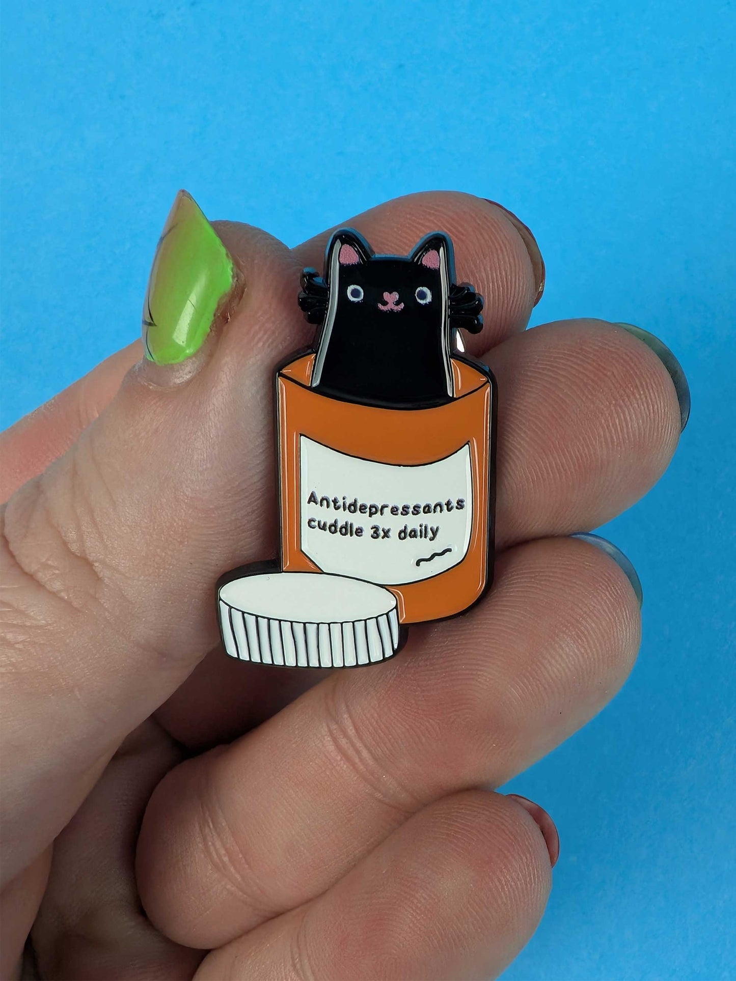 Enamel pin of a black cat in an antidepressant bottle with text, held between fingers against a blue background.