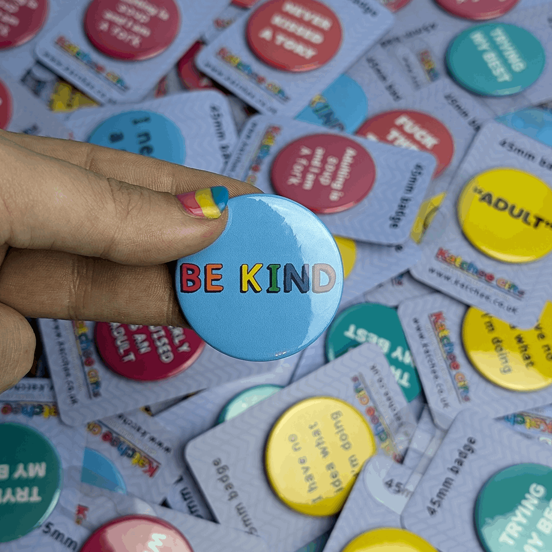 Be kind pin badge, held by a hand with nail varnish on a background of other badges.