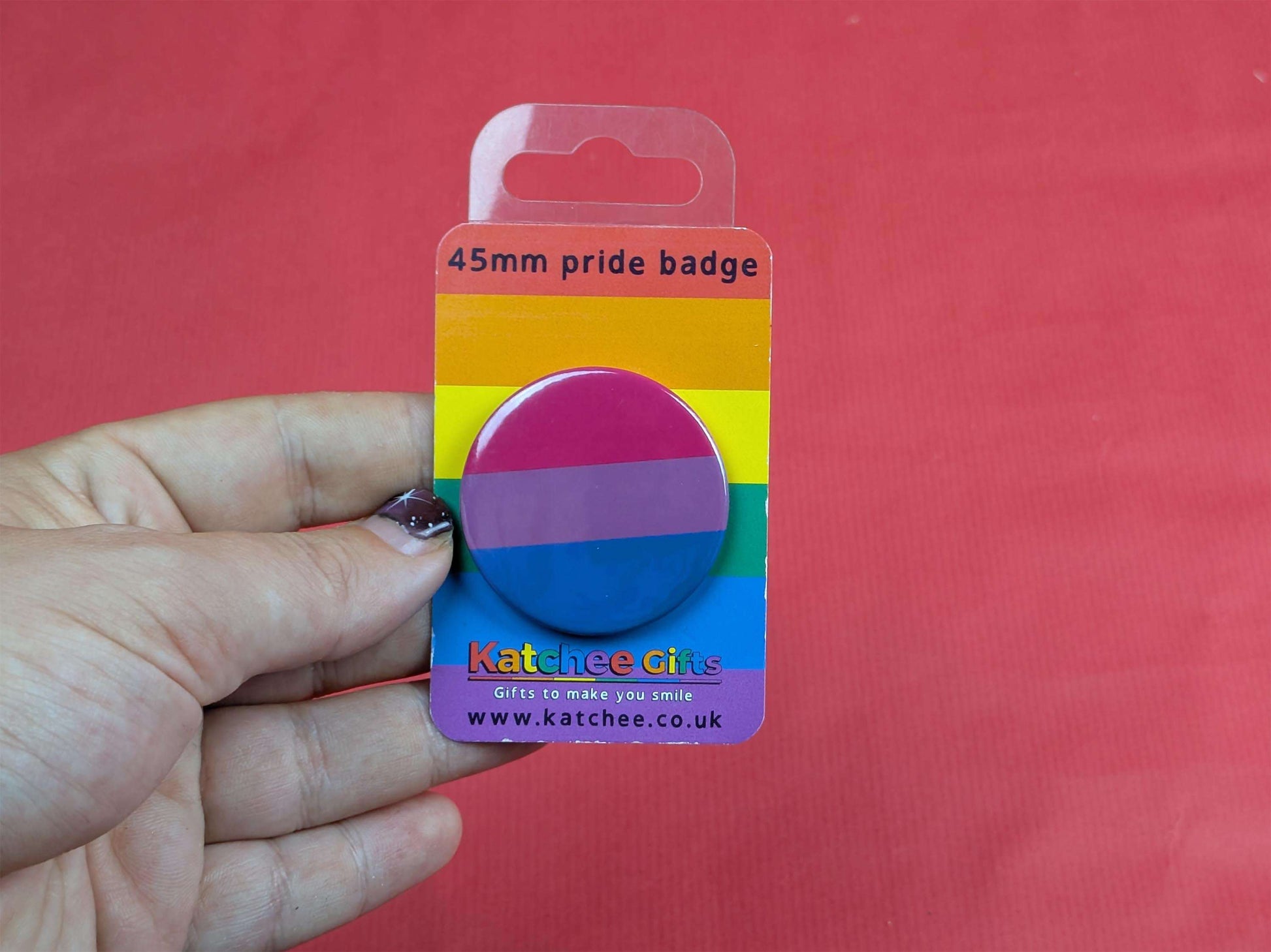 Hand holding a bisexual flag badge packaged with 'Katchee Gifts' branding on a red background.