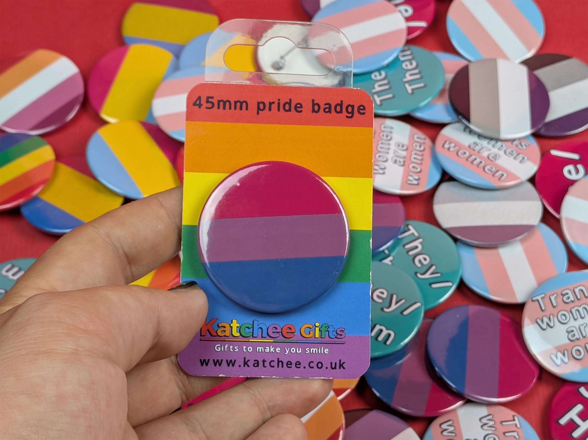 Hand holding a pride badge in packaging with a colorful background of various badges.