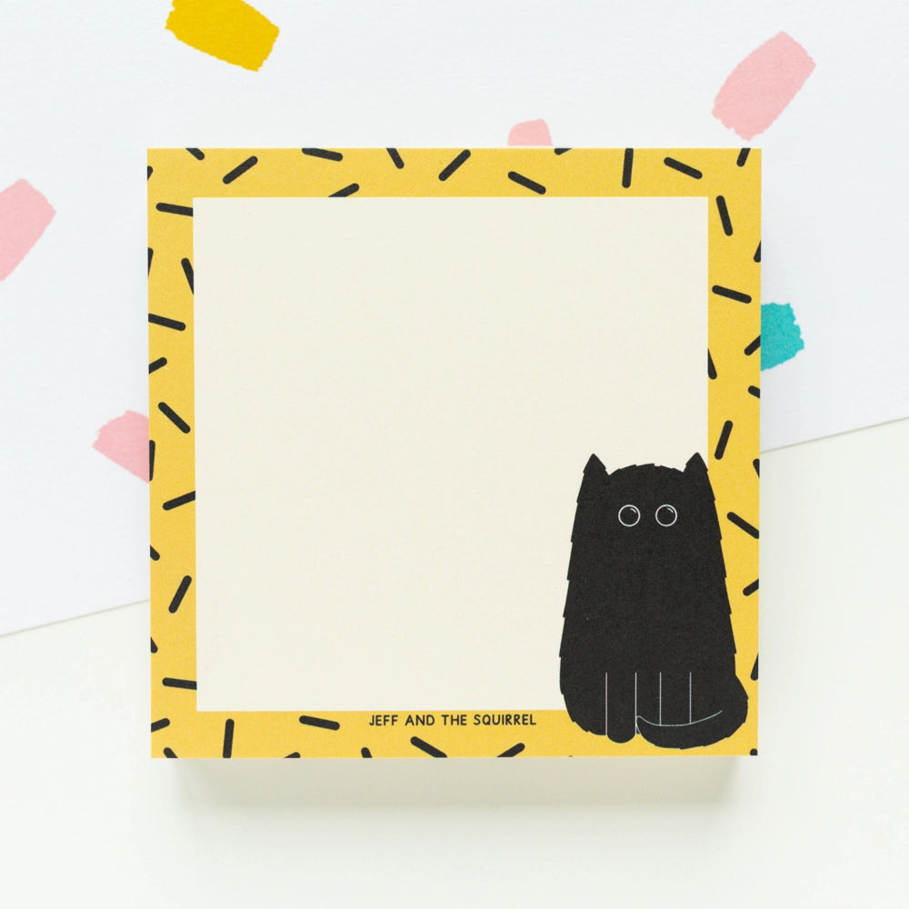 A square notepad with a yellow border patterned with black dash-like marks, a black cat illustration at the lower right, and the text 'JEFF AND THE SQUIRREL' near the bottom. The notepad is photographed on a white background with a few pastel paper confetti shapes in the corners.