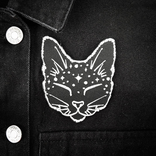 A close-up of a black denim jacket featuring a white embroidered cat face patch with stars and crescent moons on the chest.