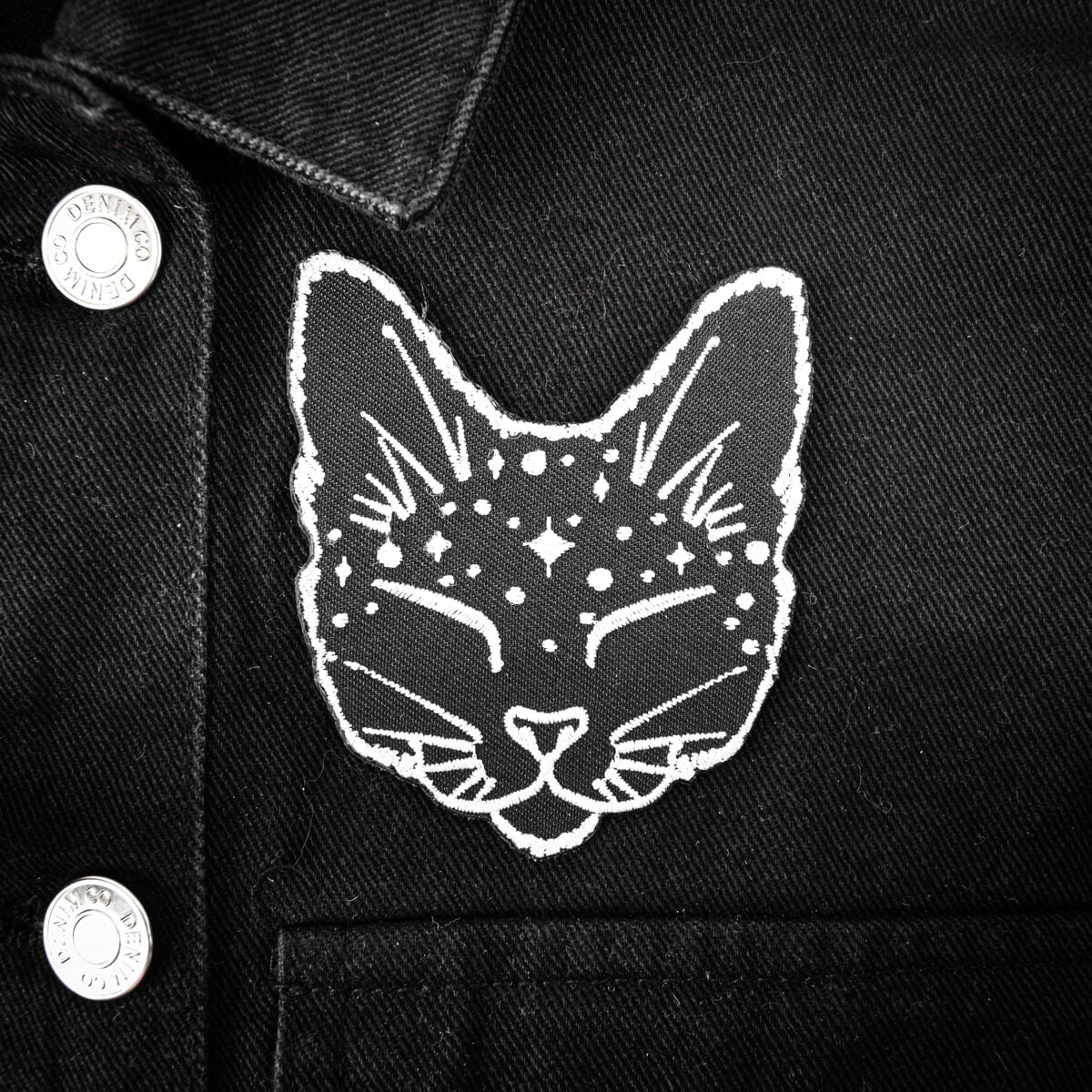 A close-up of a black denim jacket featuring a white embroidered cat face patch with stars and crescent moons on the chest.