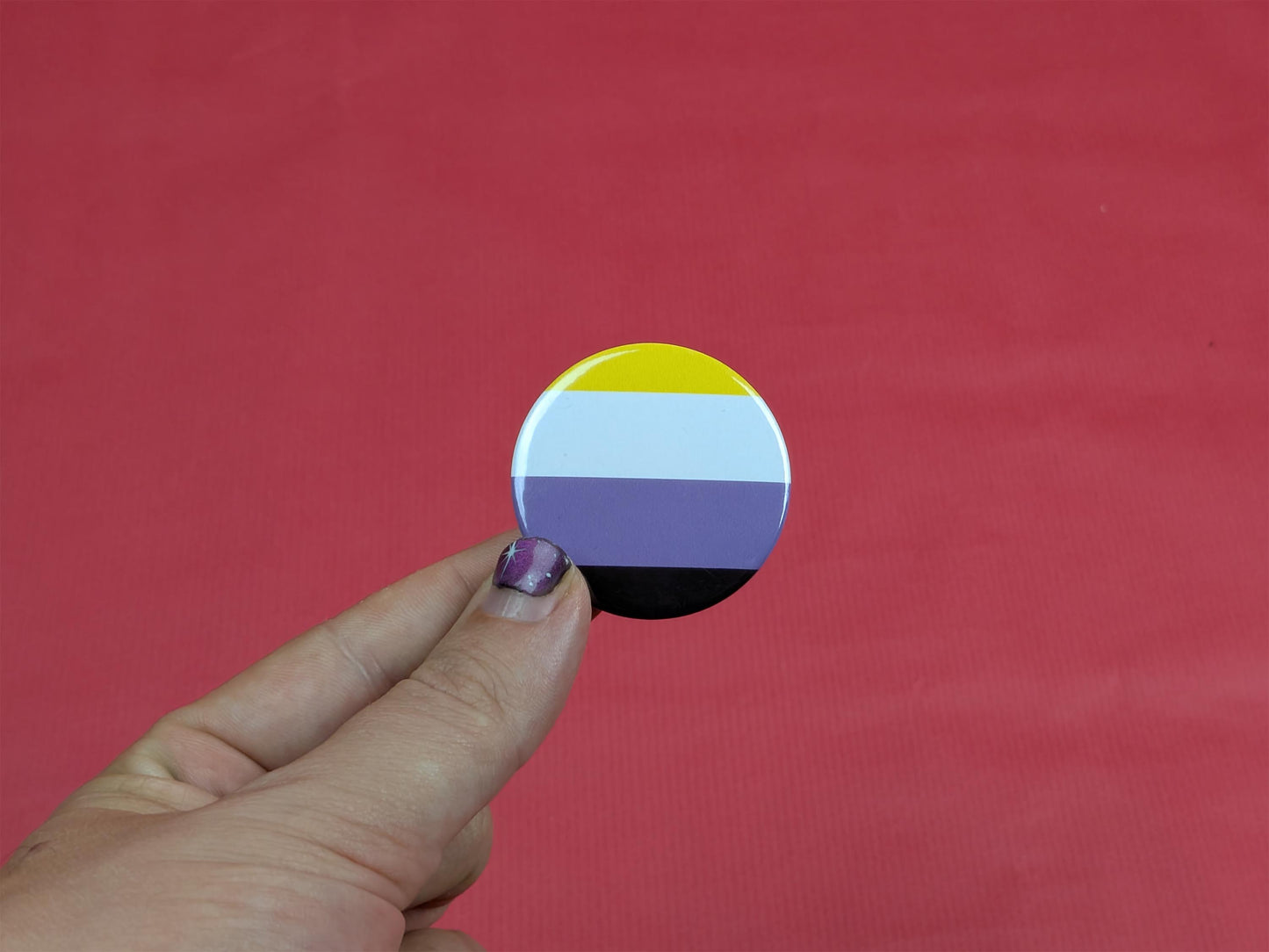 Hand holding a circular badge with a non binary flag design against a red background
