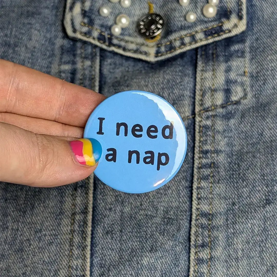 Blue button with 'I need a nap' text held by a hand against a denim background