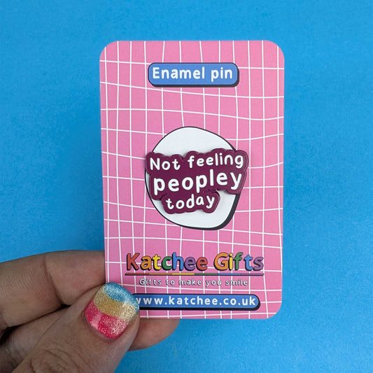 A hand holds a pink enamel pin backing card featuring a white grid pattern and a central sticker that reads 'Not feeling peopley today' in white text on a dark purple badge. The card also shows the brand name 'Katchee Gifts' and website URL at the bottom, set against a solid blue background.