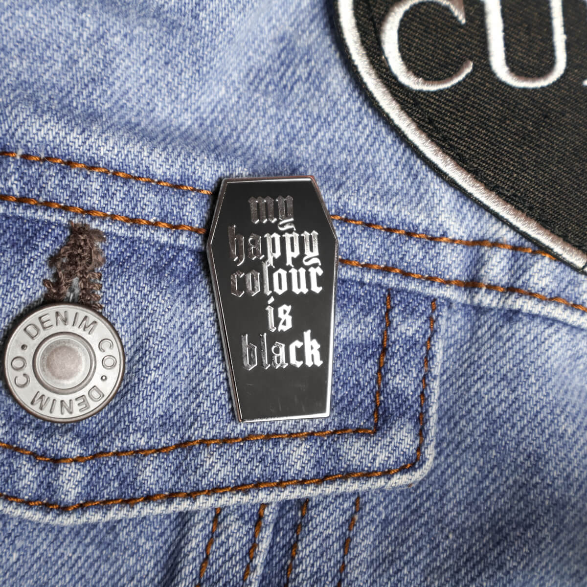 Black coffin-shaped enamel pin with 'my happy color is black' text on a denim jacket.