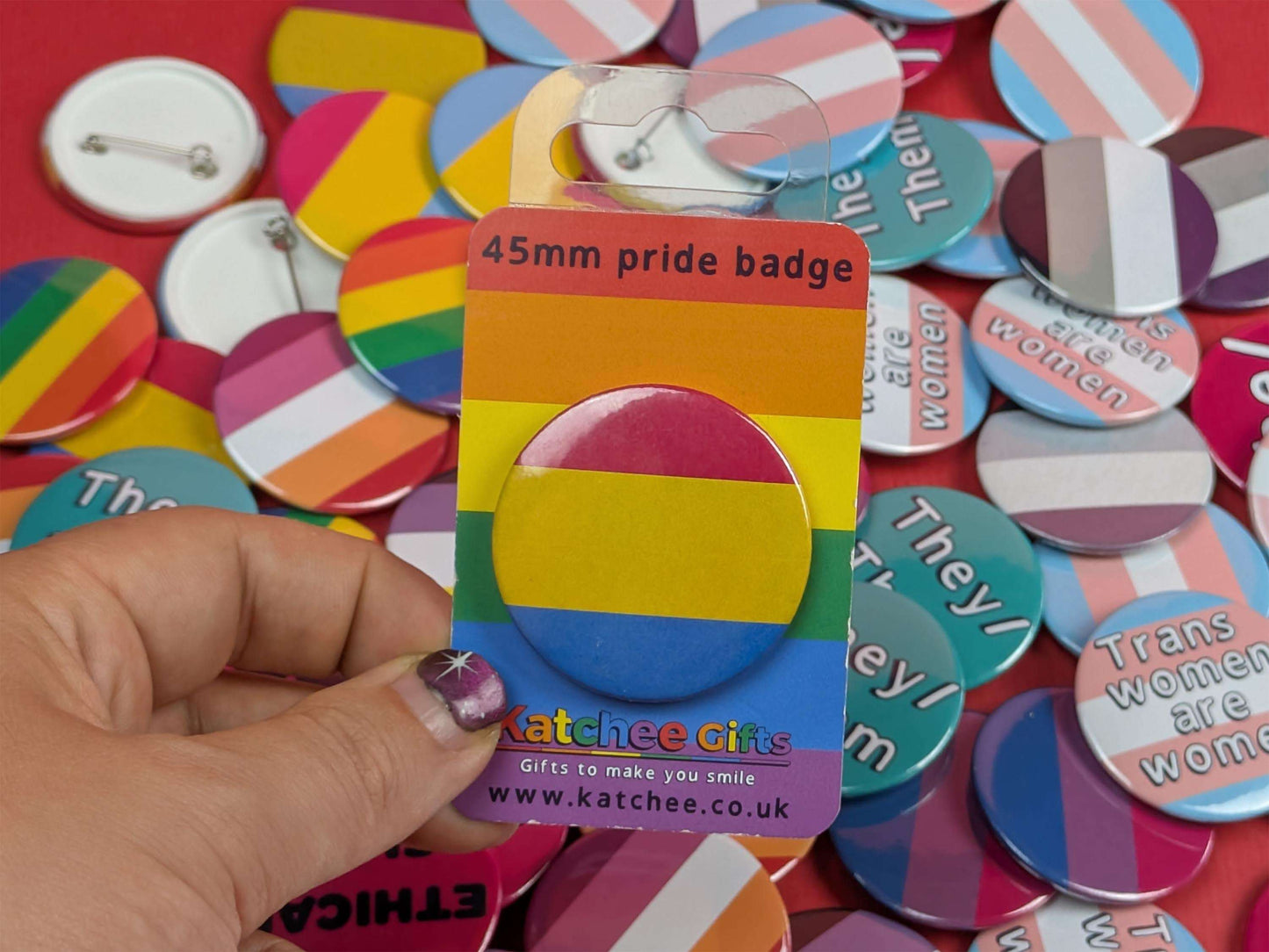Packaged 45mm pride badge with a hand holding it against a background of various colorful buttons.