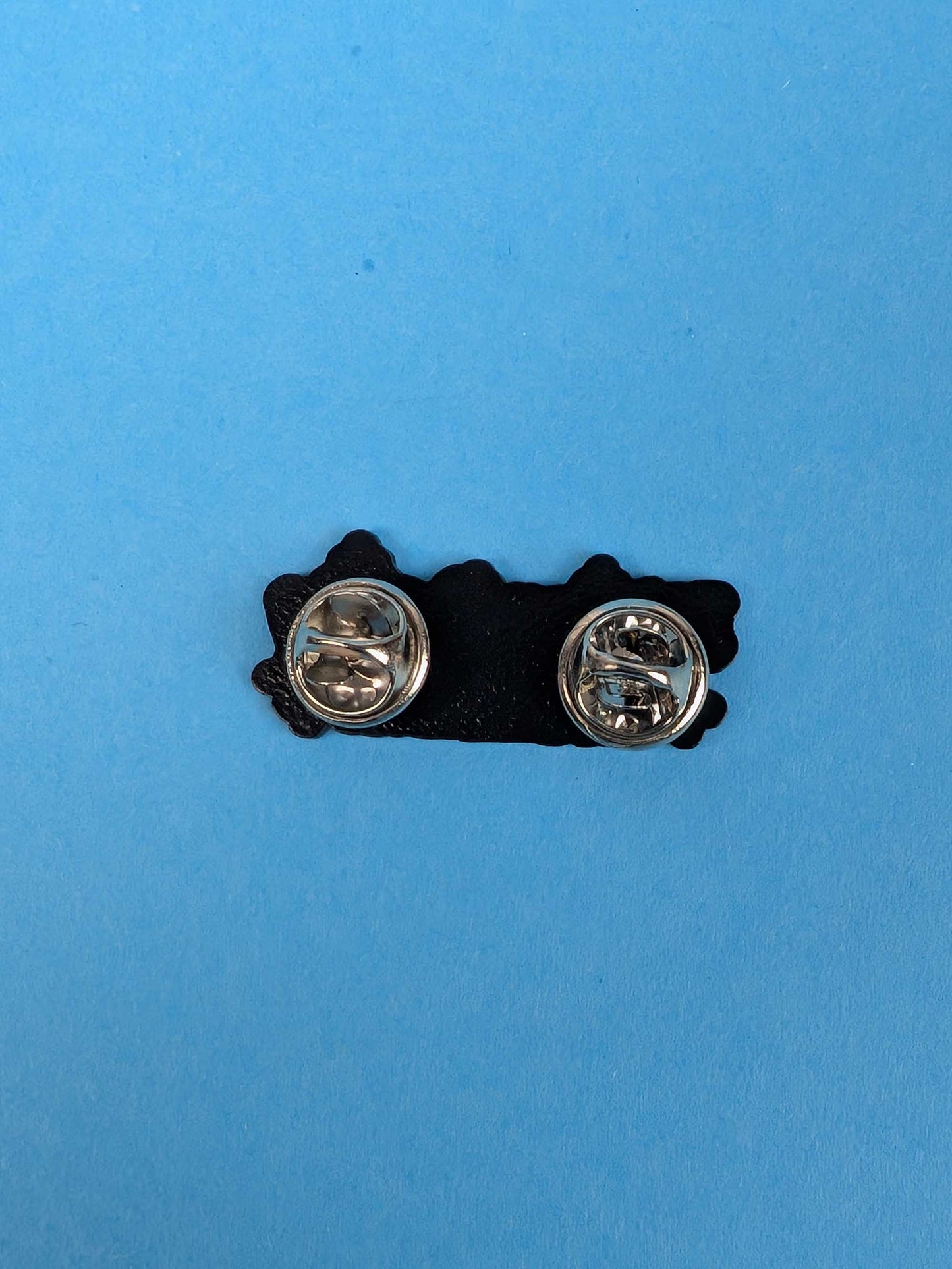 back of an enamel pin with silver clasps on a blue background