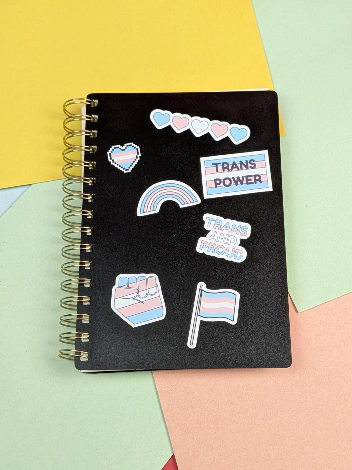 Black spiral-bound notebook with pride-themed stickers on a colorful background