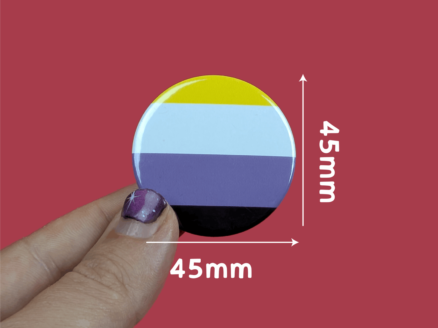 Round badge with a non binary flag design held by a hand against a red background, with measurements indicated.