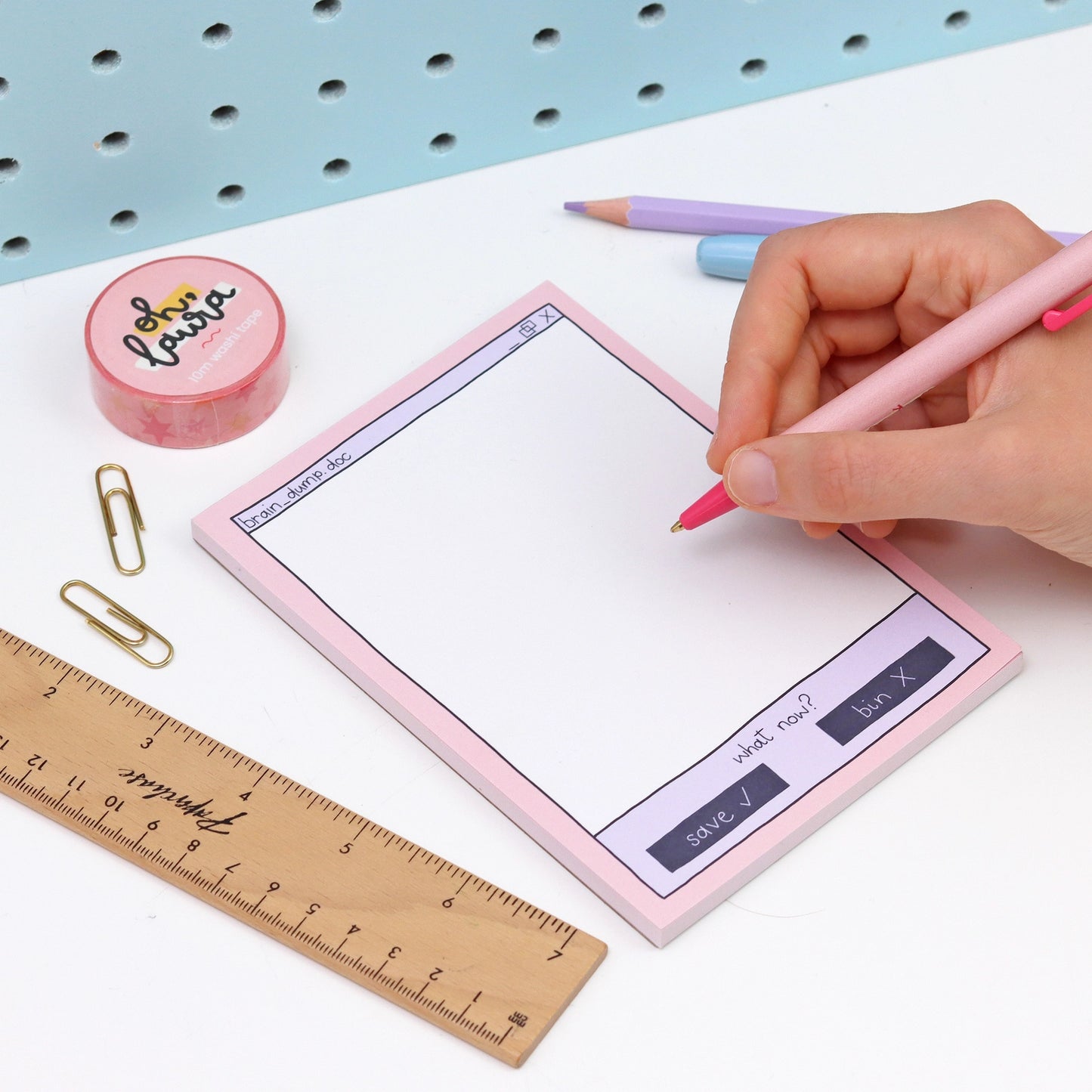 Person writing on a pink notepad with a ruler and paper clips on a white surface.