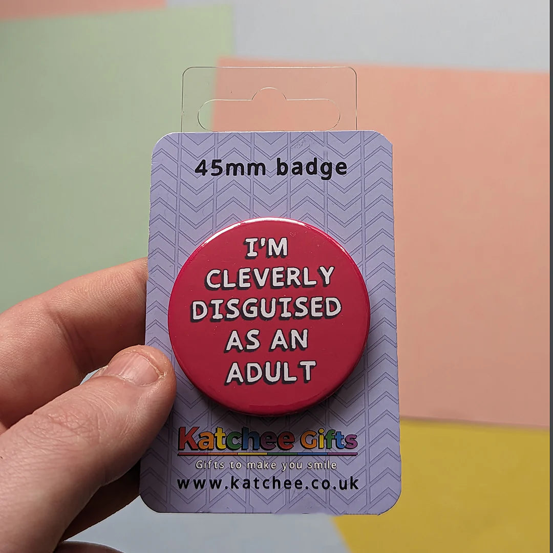 45mm badge with 'I'm cleverly disguised as an adult' text on a purple card, held by a hand against a multicolored background.
