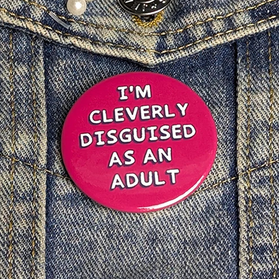 A circle pinback button which says "I'm cleverly disguised as an adult" on a denim background