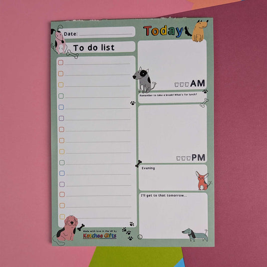 A dog-themed daily to-do list notepad with cute cartoon illustrations of various dog breeds. The pad features sections for Date, To do list, AM, PM, and Evening, decorated with colorful dog graphics on a light green background, set against a pink paper backdrop.