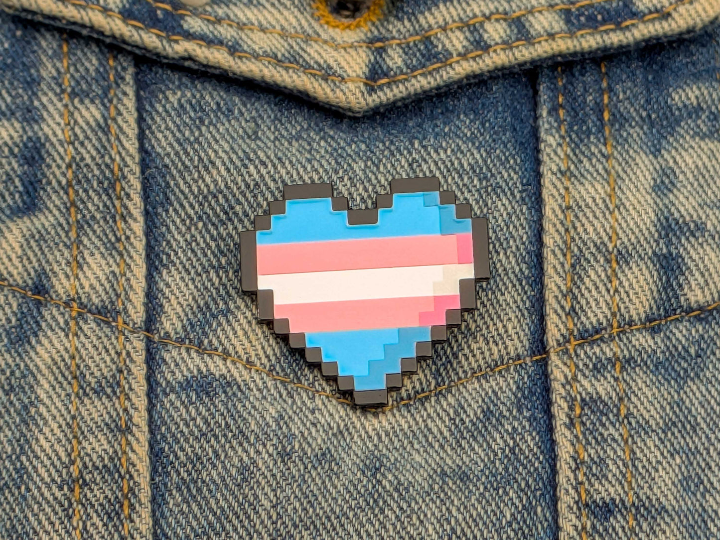 Pixelated heart-shaped pin with a transgender flag design on a denim jacket.