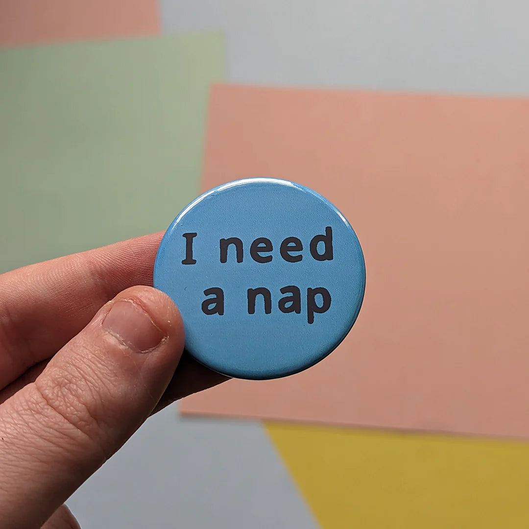 Hand holding a blue button with 'I need a nap' text against a colorful geometric background