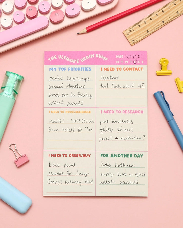 A pink and cream planner notepad titled 'The Ultimate Brain Dump' is shown in a styled flat-lay with stationery props including a pink keyboard, ruler, pencil, clips, and pens on a pink background.