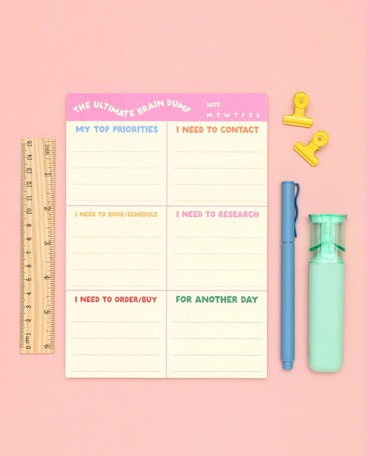 A pastel desk organizer pad titled 'THE ULTIMATE BRAIN DUMP' laid flat on a pink background, surrounded by coordinating stationery props including a wooden ruler, yellow binder clips, a blue pen, and a mint highlighter.