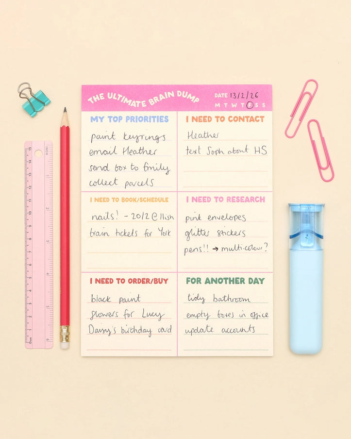 A flat-lay of a 'The Ultimate Brain Dump' notepad on a light beige background, surrounded by stationery items including a pink pencil, blue binder clip, pink ruler, pink paperclips, and a light blue water bottle.