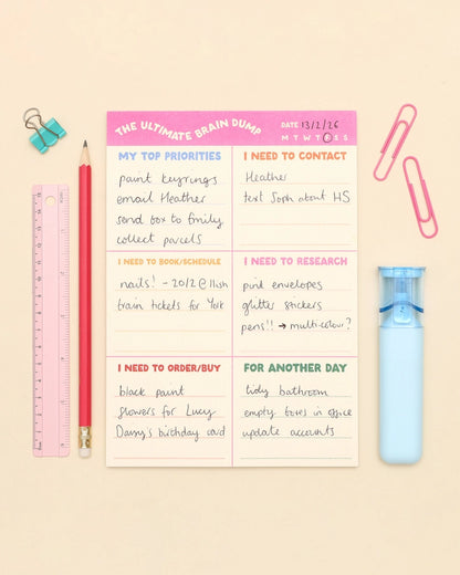 A flat-lay of a 'The Ultimate Brain Dump' notepad on a light beige background, surrounded by stationery items including a pink pencil, blue binder clip, pink ruler, pink paperclips, and a light blue water bottle.