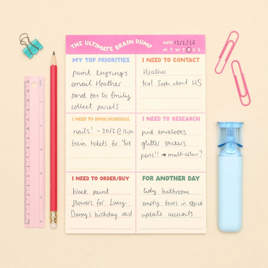A flat-lay of a 'The Ultimate Brain Dump' notepad on a beige background, surrounded by stationery props including a pink ruler, red pencil, blue binder clips, paperclips, and a light blue eraser.