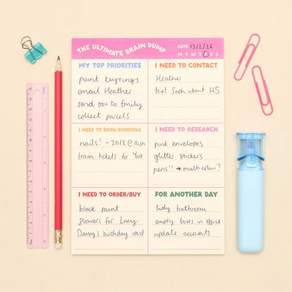 A flat-lay of a 'The Ultimate Brain Dump' notepad on a beige background, surrounded by stationery props including a pink ruler, red pencil, blue binder clips, paperclips, and a light blue eraser.