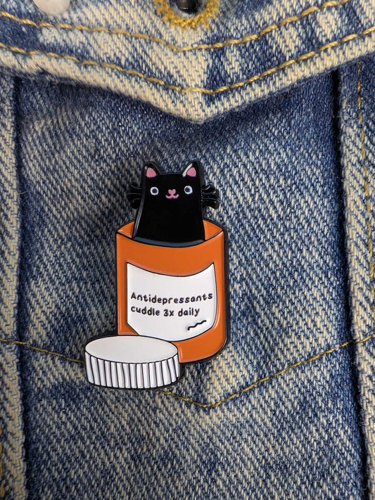 Enamel pin of a black cat in an orange antidepressants container on denim fabric