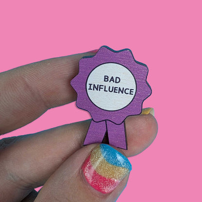 A close-up of a hand holding a purple ribbon-shaped enamel pin with a white center that reads 'BAD INFLUENCE' in black text, against a solid pink background.