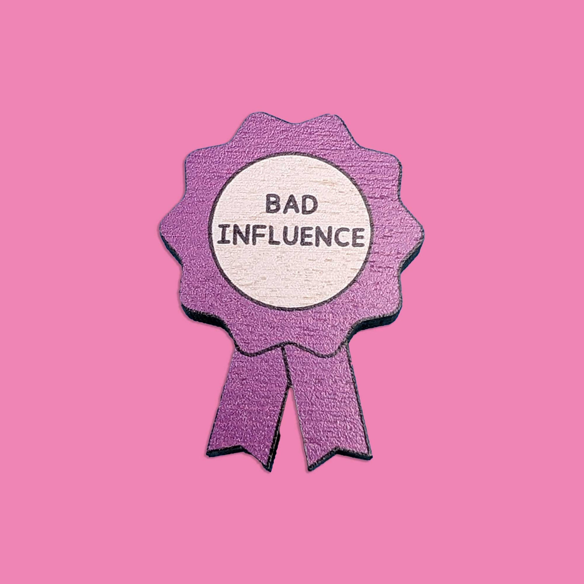 A purple ribbon pin with a white circular center reading 'BAD INFLUENCE' in black text, shown on a solid pink background.