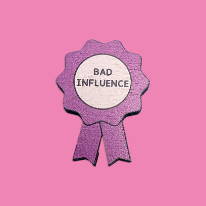 A purple ribbon pin with a white circular center reading 'BAD INFLUENCE' in black text, shown on a solid pink background.