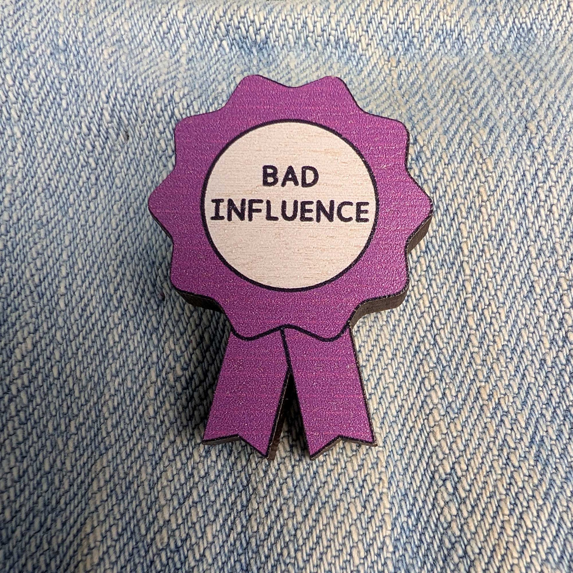 A purple ribbon-shaped enamel pin with a white center reading 'BAD INFLUENCE' in black text, placed on a blue denim background.