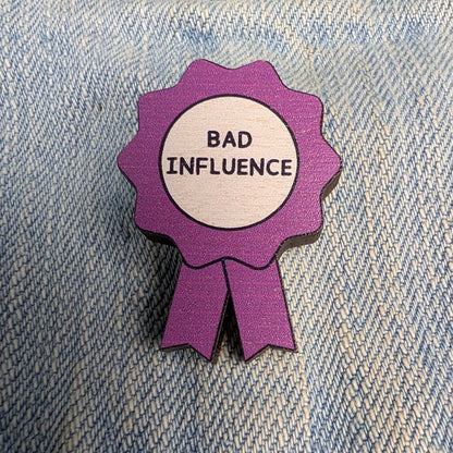 A purple ribbon-shaped enamel pin with a white center reading 'BAD INFLUENCE' in black text, placed on a blue denim background.