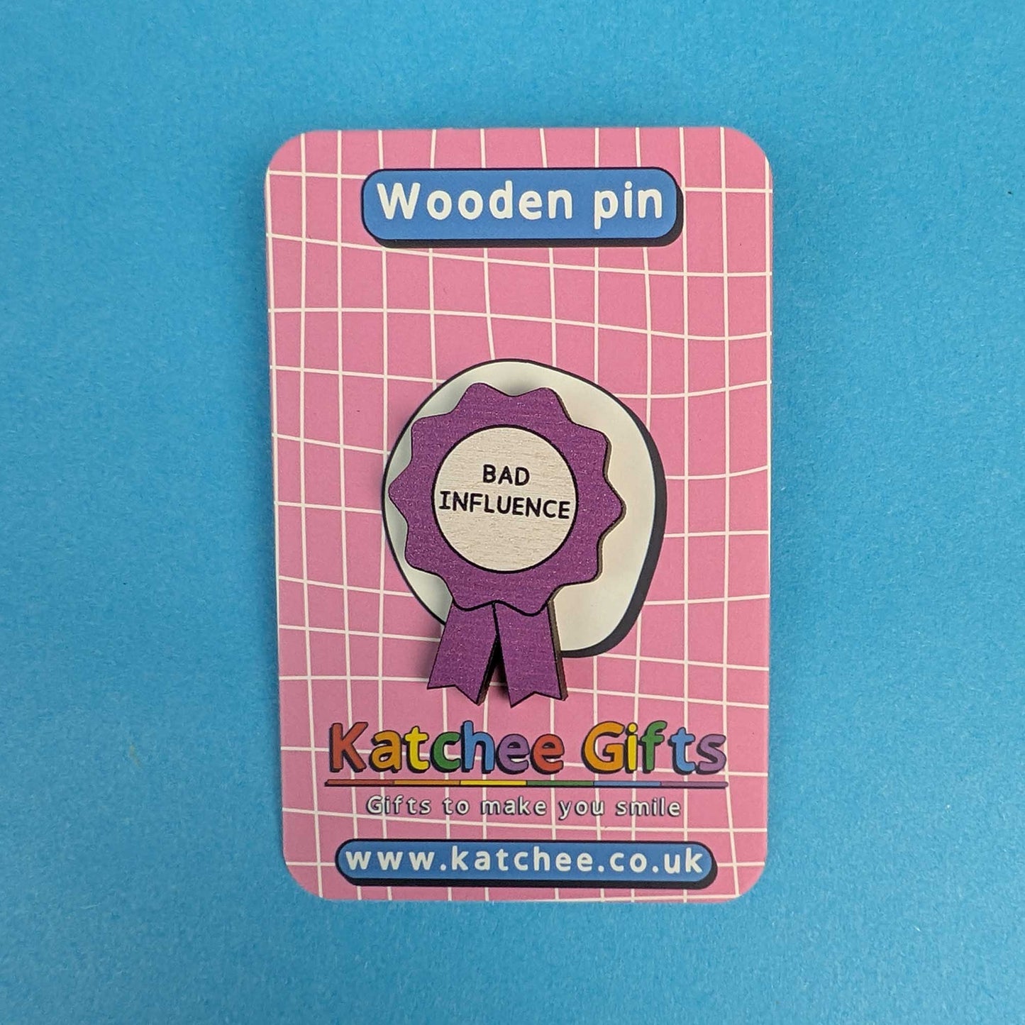 A wooden pin shaped like a purple and white award ribbon with the words 'BAD INFLUENCE' in black, mounted on a pink card labeled 'Wooden pin' and branded 'Katchee Gifts'.