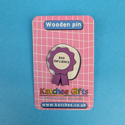 A wooden pin shaped like a purple and white award ribbon with the words 'BAD INFLUENCE' in black, mounted on a pink card labeled 'Wooden pin' and branded 'Katchee Gifts'.