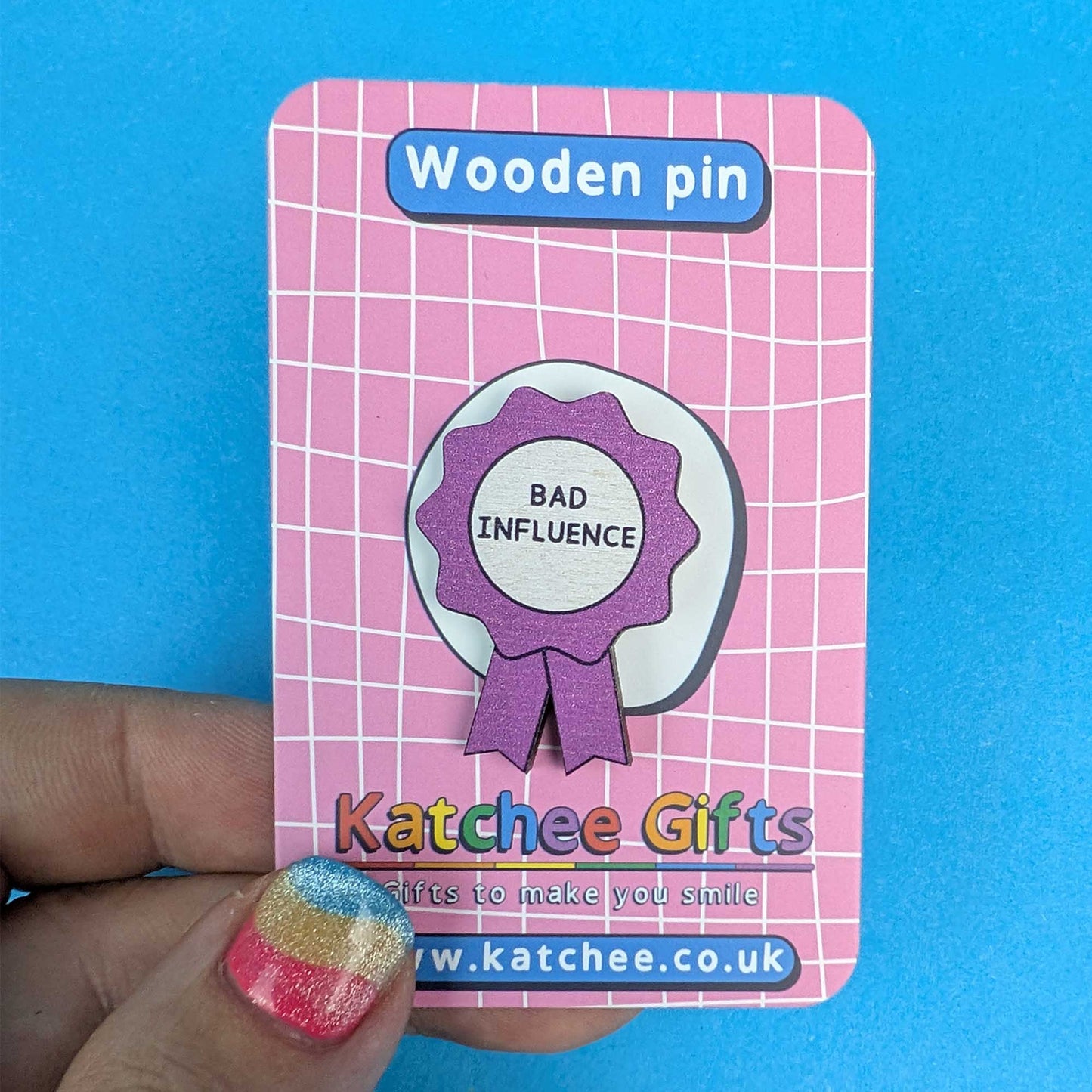 A wooden pin shaped like a purple and white award ribbon badge with the text 'BAD INFLUENCE', mounted on a pink card labeled 'Wooden pin' and branded 'Katchee Gifts'. A hand with glittery nail polish holds the card against a bright blue background.