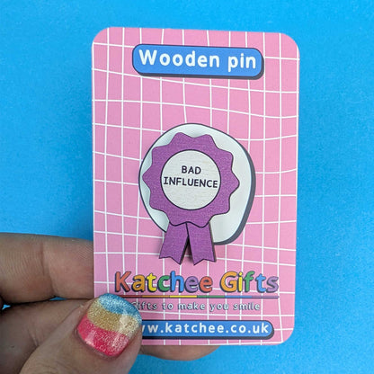 A wooden pin shaped like a purple and white award ribbon badge with the text 'BAD INFLUENCE', mounted on a pink card labeled 'Wooden pin' and branded 'Katchee Gifts'. A hand with glittery nail polish holds the card against a bright blue background.