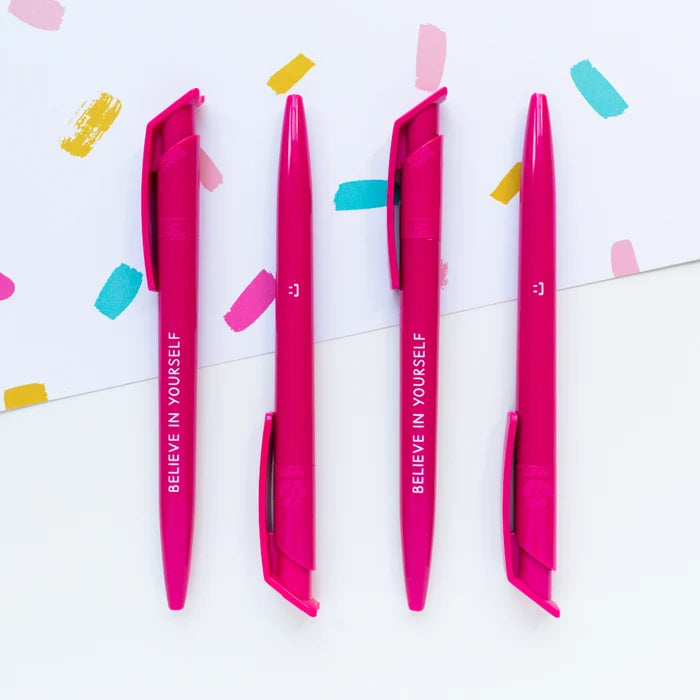 Four bright pink retractable gel pens with white text reading 'BELIEVE IN YOURSELF' arranged vertically on a white background with colorful paint strokes and confetti.