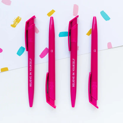 Four bright pink retractable gel pens with white text reading 'BELIEVE IN YOURSELF' arranged vertically on a white background with colorful paint strokes and confetti.
