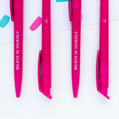 Four bright pink retractable pens with white text reading 'BELIEVE IN YOURSELF' and a smiley face symbol, arranged vertically on a white background with colorful paint strokes.