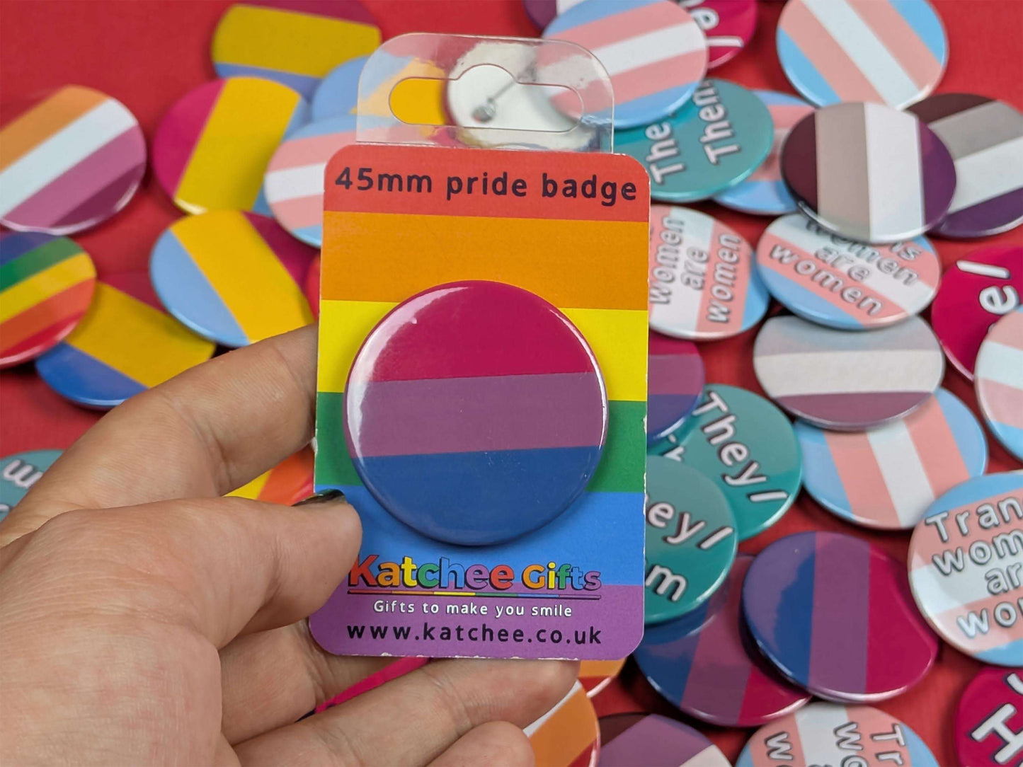 Hand holding a pride badge in packaging with a colorful background of various badges.