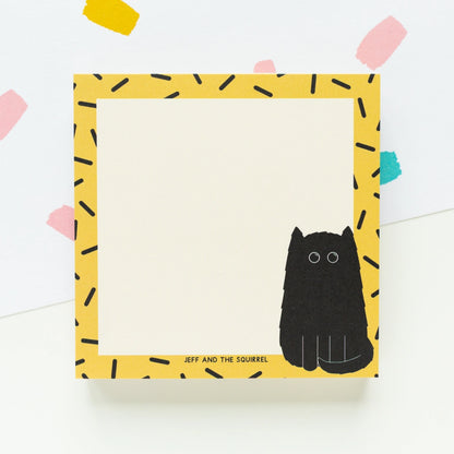A square notepad with a yellow border patterned with black dash-like marks, a black cat illustration at the lower right, and the text 'JEFF AND THE SQUIRREL' near the bottom. The notepad is photographed on a white background with a few pastel paper confetti shapes in the corners.