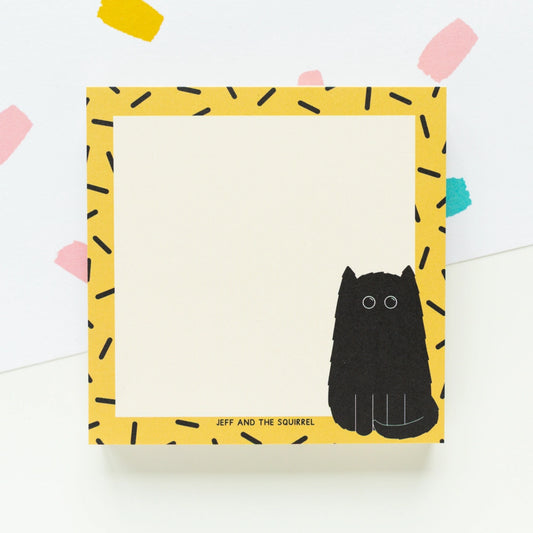 A square notepad with a yellow border patterned with black dash-like marks, a black cat illustration at the lower right, and the text 'JEFF AND THE SQUIRREL' near the bottom. The notepad is photographed on a white background with a few pastel paper confetti shapes in the corners.
