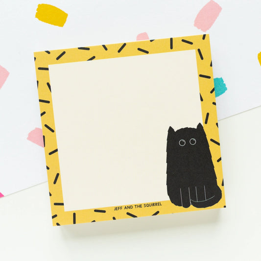 A square notepad with a yellow border patterned with black dash-like marks and a black squirrel illustration in the lower right corner, photographed on a white background with colorful confetti accents.