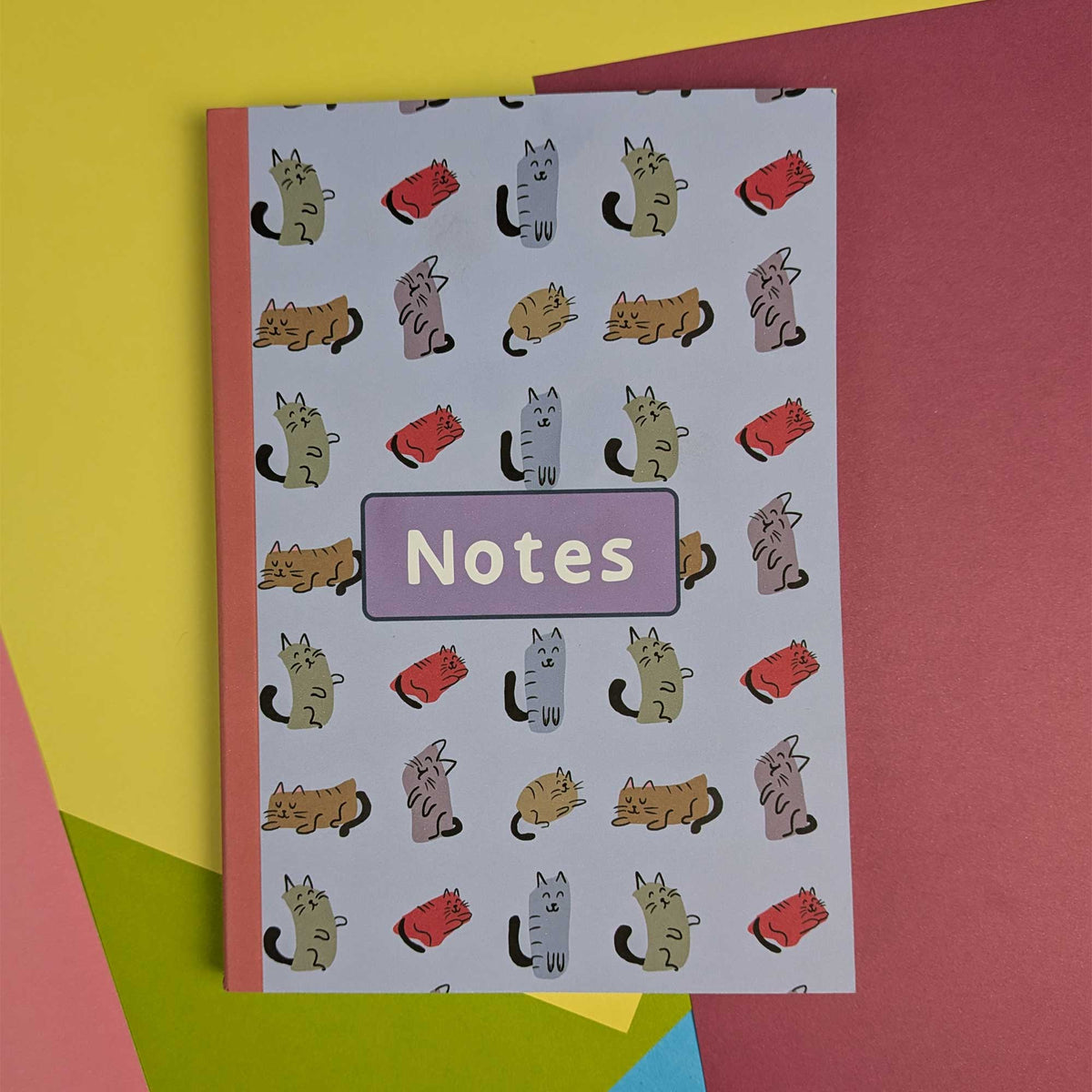 A notebook with a white background covered in a repeating pattern of cartoon cats in different colors, with a lavender label reading 'Notes' in the center. It is photographed on top of bright colored paper sheets.