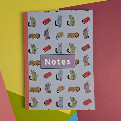 A notebook with a white background covered in a repeating pattern of cartoon cats in different colors, with a lavender label reading 'Notes' in the center. It is photographed on top of bright colored paper sheets.