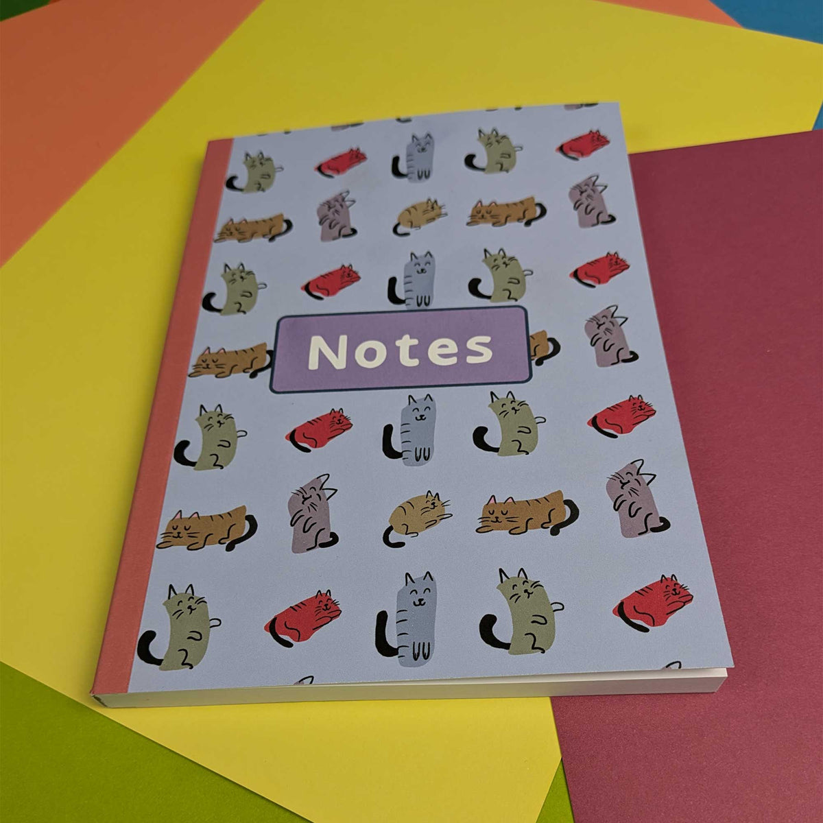 A notebook with a light lavender cover decorated with a repeating pattern of cartoon cats in various colors, featuring a purple label that reads 'Notes'. The notebook is placed on a colorful background of overlapping paper sheets in yellow, orange, pink, and green.