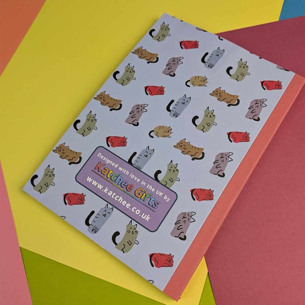 A notebook with a light lavender cover printed with a repeating pattern of cartoon cats in various colors, resting on a colorful background of yellow, pink, and green paper. The brand 'Katchee Gifts' is clearly visible on the purple label on the cover.