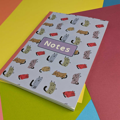 A notebook with a white cover decorated with a repeating pattern of illustrated cats in various colors and a purple label reading 'Notes', photographed on top of bright colored paper sheets.