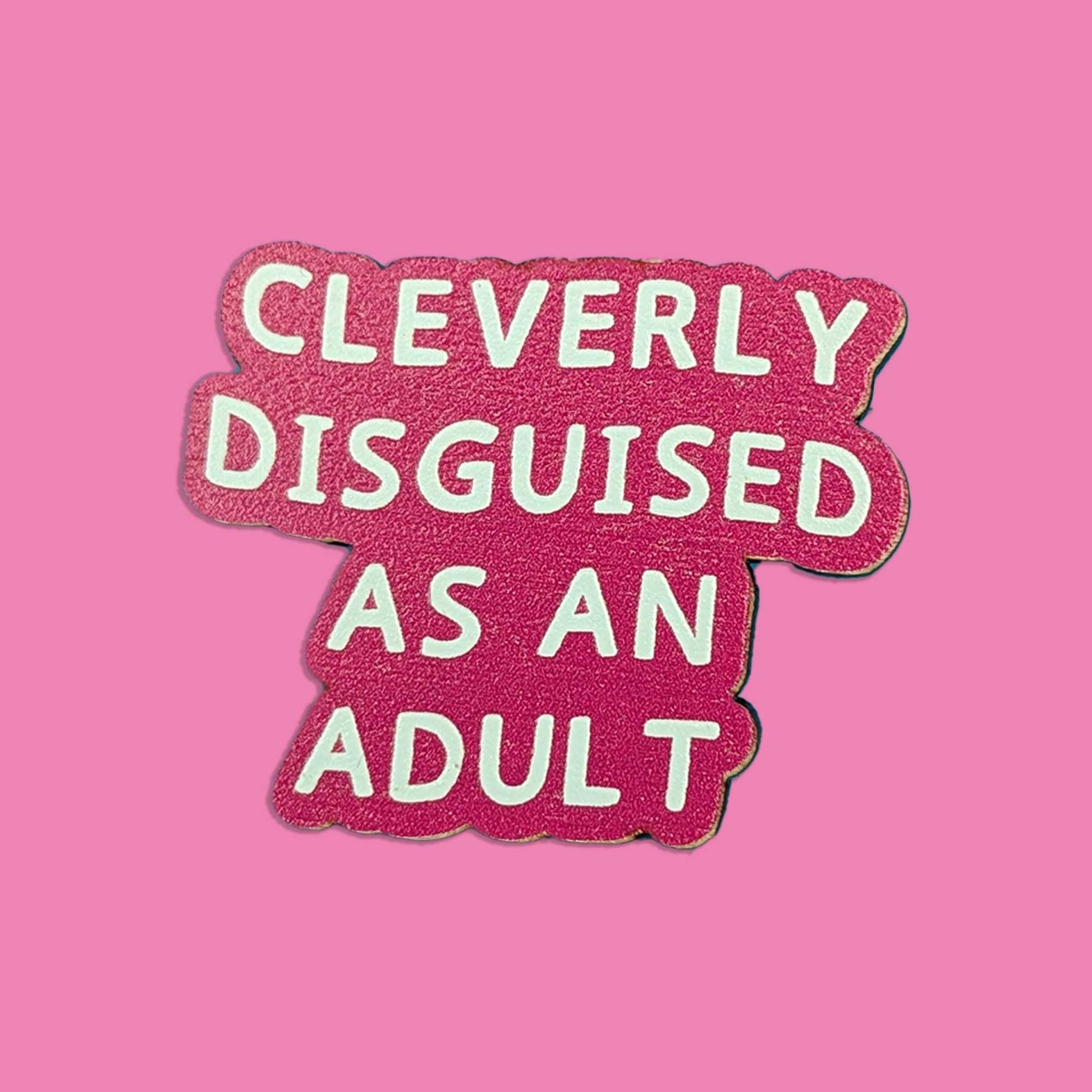 A pink glittery enamel pin with white text reading 'CLEVERLY DISGUISED AS AN ADULT' on a solid pink background.